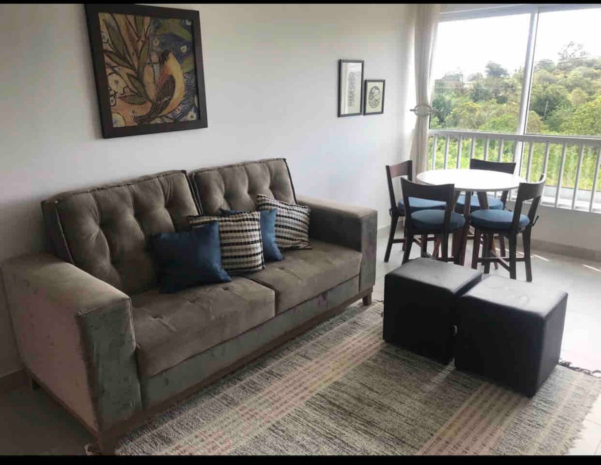 A comfortable living area features a light-colored sofa adorned with blue accent pillows. A small dining table with four chairs sits beside a large window, providing views of the surrounding greenery. A textured rug adds warmth to the space, complemented by framed artwork on the wall.
