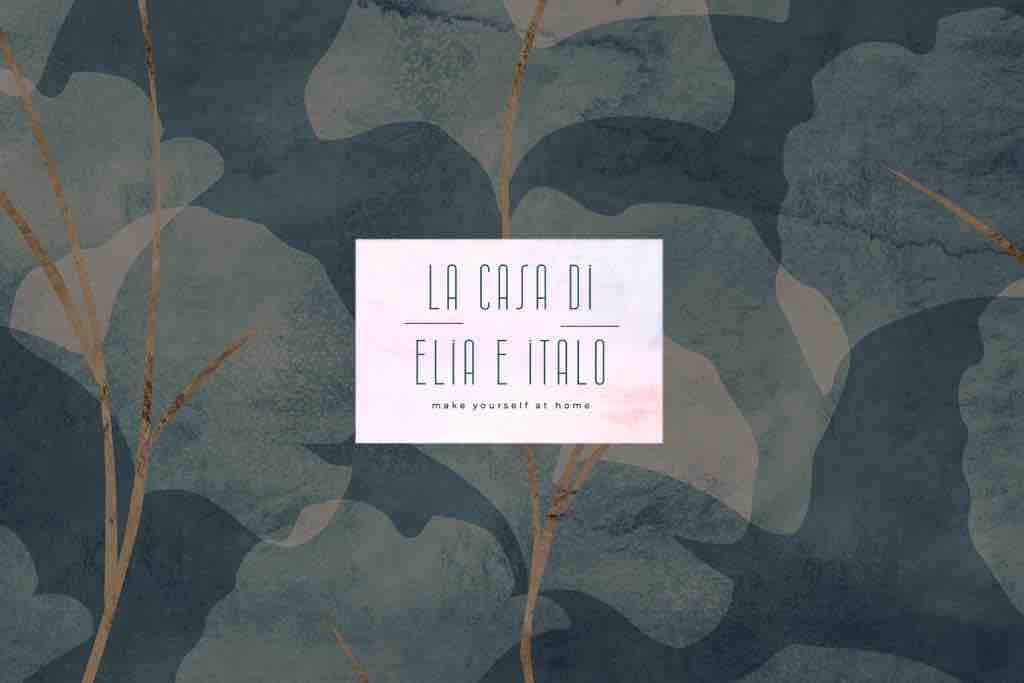 A decorative wall display features a blend of soft green and blue hues, adorned with abstract leaf patterns. The center showcases a welcoming sign, reading 'LA CASA DI ELIA E ITALÒ' with a subtle message inviting guests to feel at home.