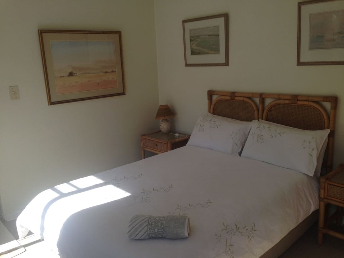 A calming bedroom featuring a spacious bed with light-colored bedding and decorative pillows. Twin bedside tables are positioned on either side, each adorned with a lamp. Artwork hangs on the walls, and sunlight streams in, creating a serene and inviting environment.