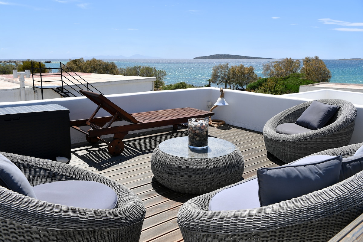 A rooftop deck features comfortable woven seating arrangements, a round table, and a sunbed. The sea and distant coastline provide a serene backdrop under a clear sky, enhancing the space's inviting atmosphere for relaxation.