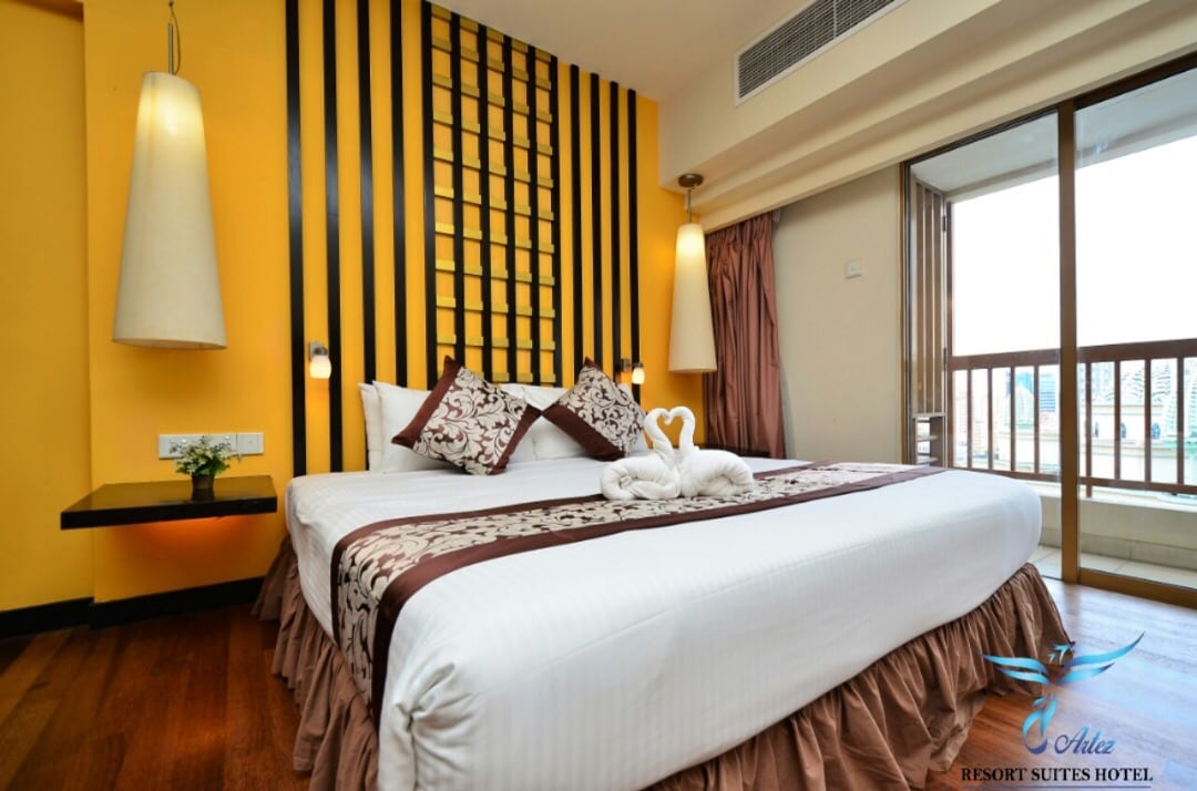 A spacious room features a king-sized bed adorned with elegant decorative pillows and a towel sculpture. A vibrant yellow accent wall is complemented by a grid-patterned design. Natural light filters in through a large window leading to a private balcony.