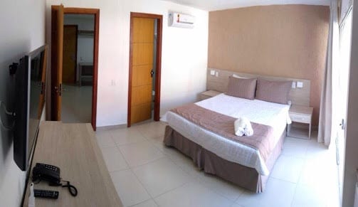 A well-lit bedroom features a spacious bed with neutral-toned bedding and decorative pillows. Two nightstands flank the bed, and a television is mounted on the wall opposite. The room includes a subtle color palette and a window that allows natural light to enter.