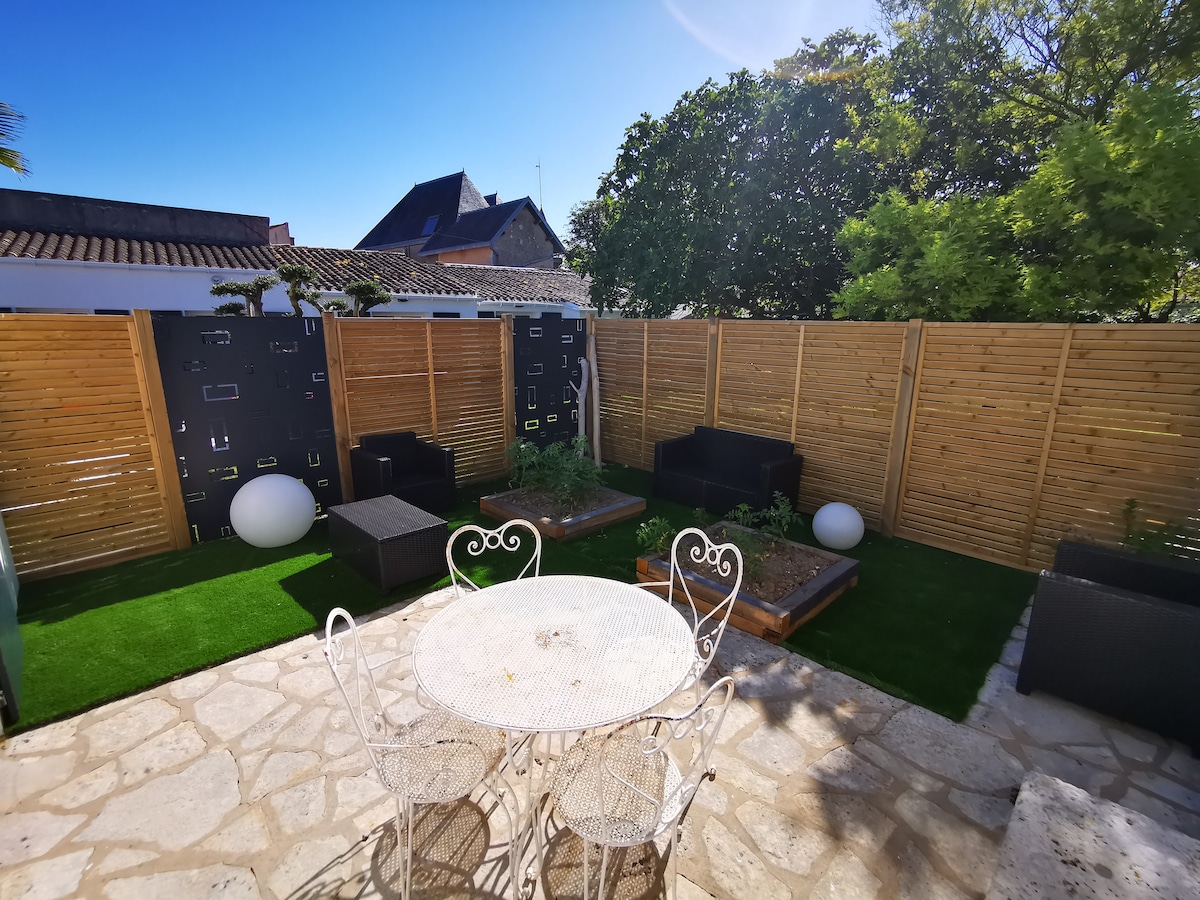 An outdoor garden area features a circular table surrounded by four white chairs. Two dark seating arrangements with cushions are positioned on green grass, while wooden screens provide privacy. A landscaped garden with plants is visible, enhanced by decorative lighting elements.