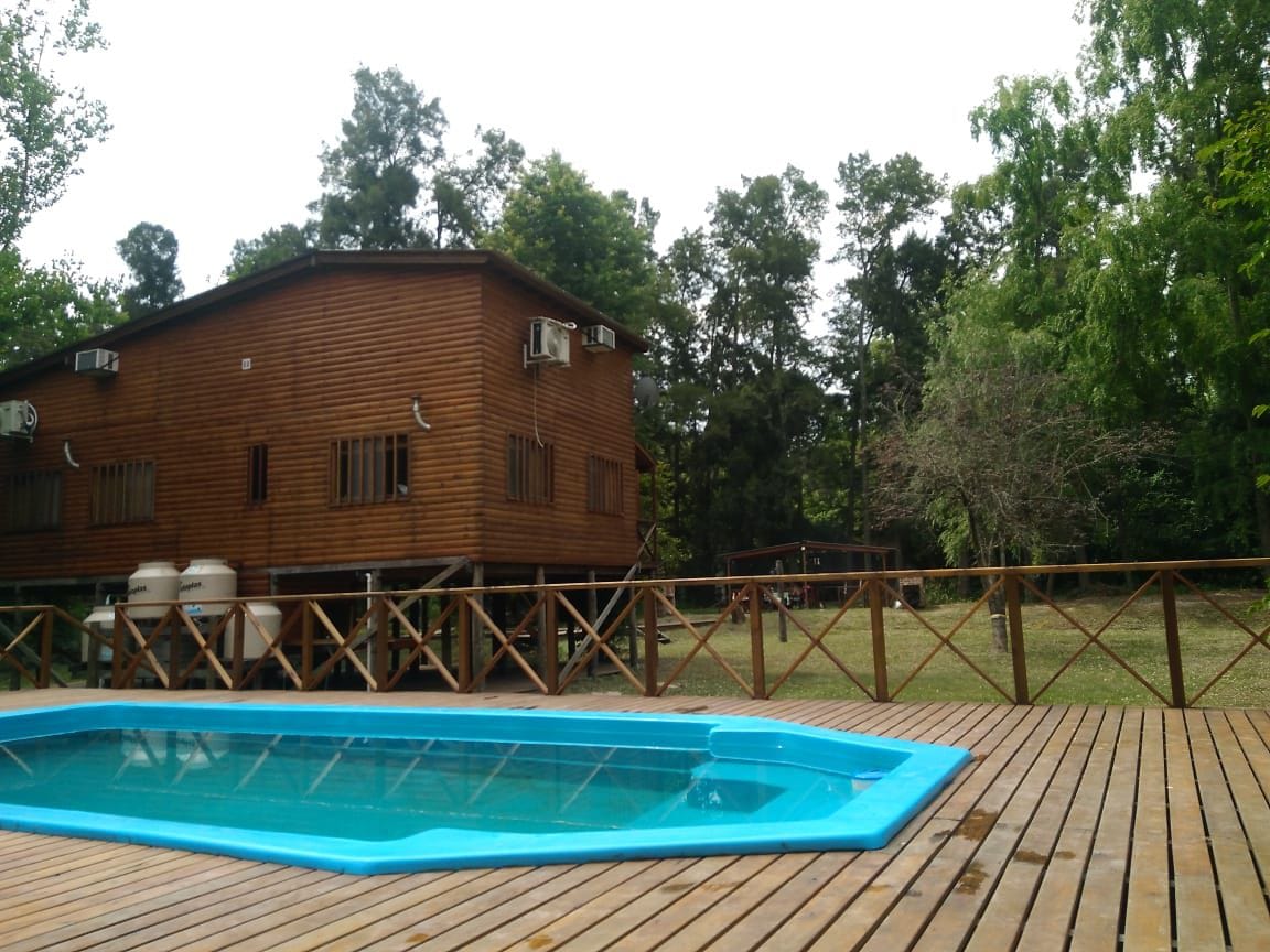 Two wooden cabins are situated amidst lush greenery, with air conditioning units visible on the exterior. A turquoise pool is framed by a wooden deck, providing a refreshing outdoor space for relaxation. The natural surroundings enhance the serene atmosphere.