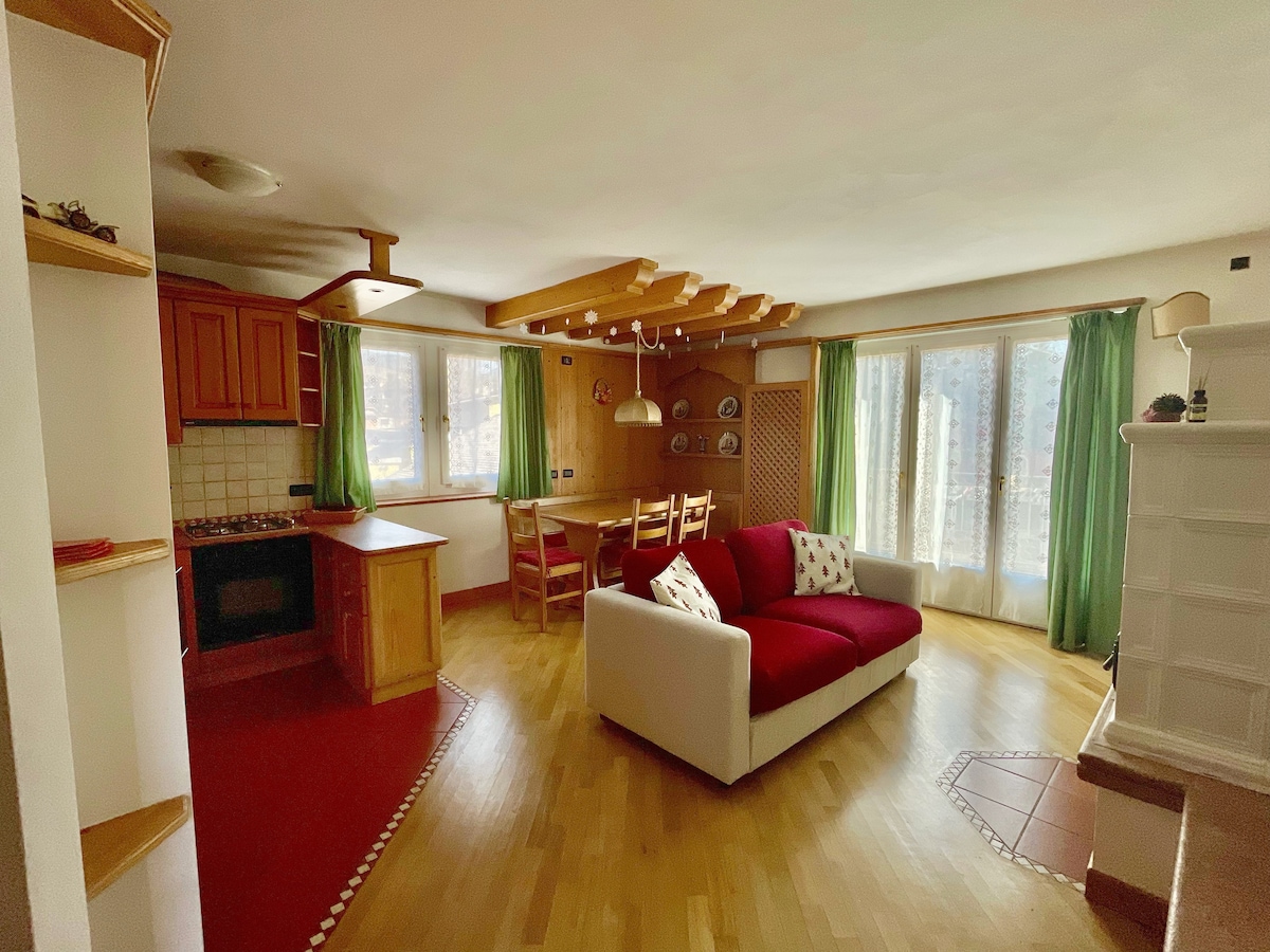A bright living area features a red sofa and a wooden dining table surrounded by four chairs. Natural light floods the space through large windows, which are dressed in green curtains. The kitchen area is visible, showcasing wooden cabinetry and appliances.