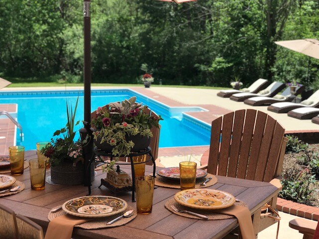 A dining table is set for outdoor meals, featuring decorative plates and glasses. In the background, a clear swimming pool is surrounded by lounge chairs and greenery, creating a serene environment for relaxation.