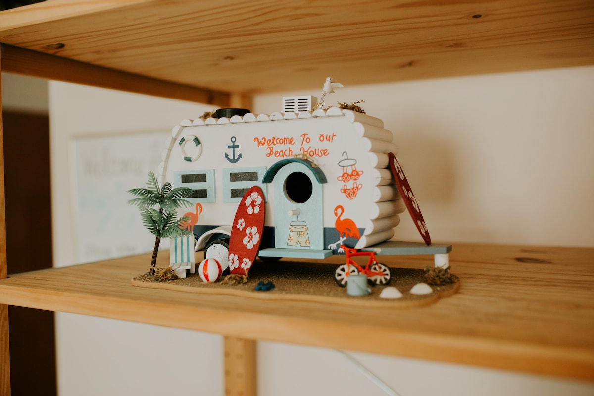 A decorative beach house model sits on a wooden shelf. The bright, whimsical design features a white exterior with colorful accents, including a flamingo, surfboards, and beach-themed decorations. Small details like palm trees and beach balls enhance the playful representation of a beachside retreat.