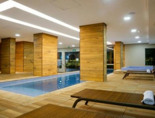 An indoor swimming area features a narrow pool bordered by wooden columns. Sun loungers are placed alongside the pool. Natural light filters in through large windows, creating a bright and airy ambiance. A whirlpool is visible at one end of the space.