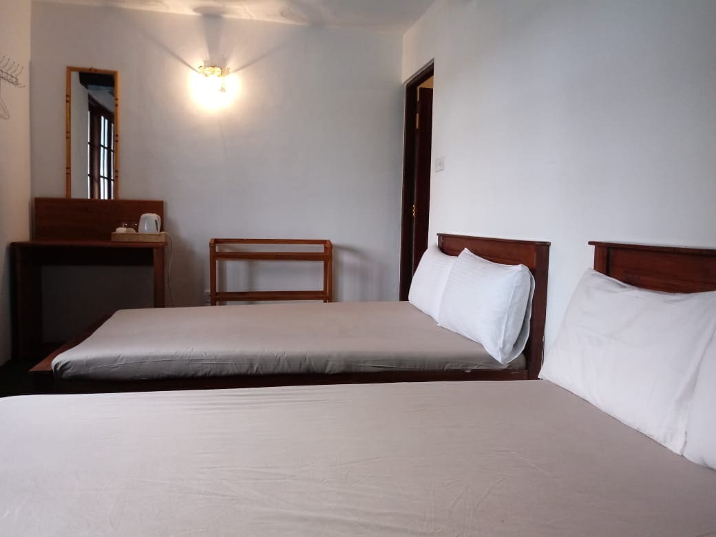 A simple and functional sleeping area is featured, containing two spacious beds with neutral-toned bedding. A wooden desk and stool are positioned nearby, alongside a mirror and an electric kettle. Natural light enters through a window, illuminating the room's clean, white walls.