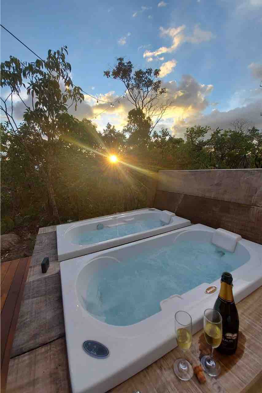 Two stylish hot tubs are situated on a wooden deck, surrounded by lush greenery. A sunset casts a warm glow over the scene, with glasses of champagne placed nearby, suggesting a serene atmosphere for relaxation.