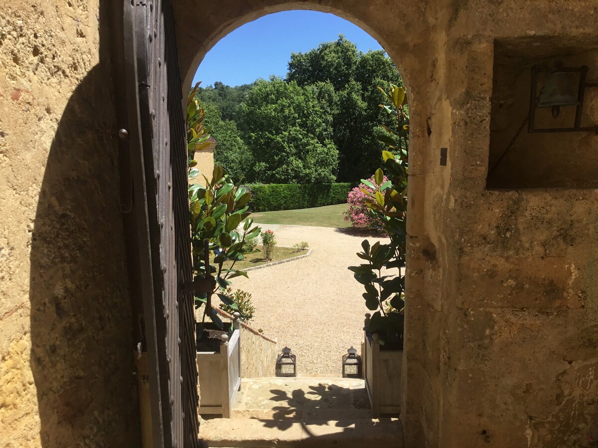 An arched doorway opens to a scenic view of the garden, framed by lush greenery. Planters with vibrant plants and lanterns are positioned at the base of the entrance, while a pebbled pathway leads through the landscaped grounds.