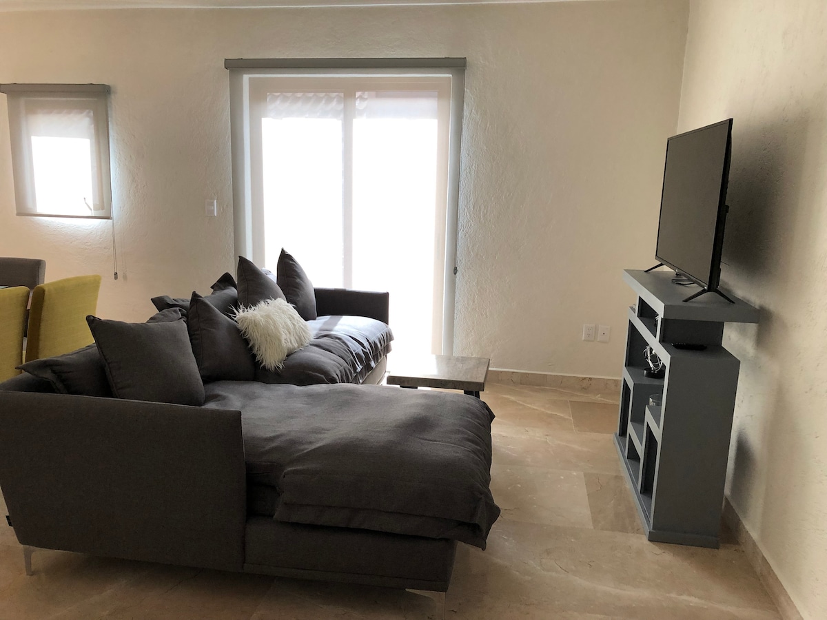 A comfortable living area is presented, featuring a spacious gray sectional sofa adorned with throw pillows. A small table sits beside the sofa, and a flat-screen television is mounted on a light gray media console. Natural light filters through the sliding glass doors.