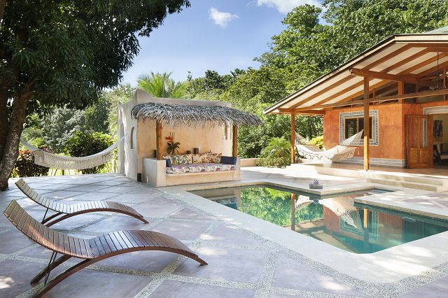 An inviting outdoor area features a serene pool surrounded by a tiled patio and wooden lounge chairs. A thatched-roof seating area offers a cozy spot for relaxation, with lush greenery framing the space. Hammocks hang nearby, enhancing the connection with nature.