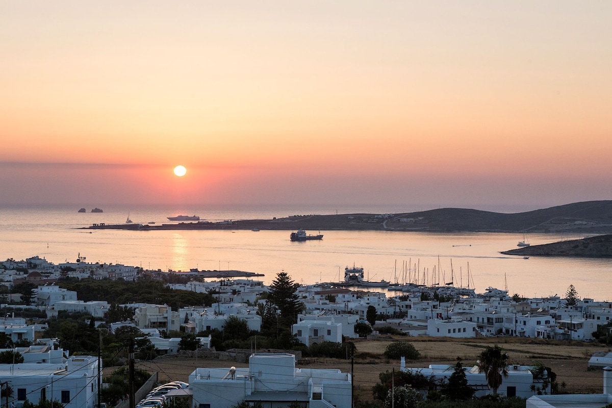 The image captures a serene sunset over the Aegean Sea, with a gentle gradient of orange and pink hues in the sky. In the foreground, white buildings are surrounded by lush greenery, while boats can be seen gently bobbing in the water.