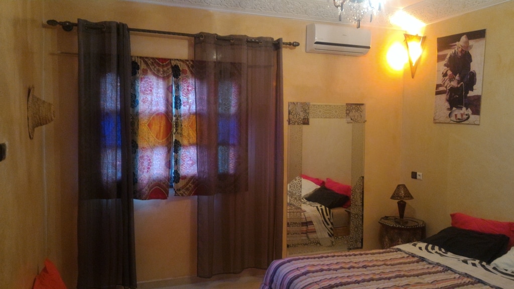 A cozy bedroom features warm, earthy tones and patterned curtains filtering natural light. A mirror is placed near the window, reflecting a tastefully decorated space. A spacious bed is adorned with colorful linens, and soft textures are present in the ambient decorations.