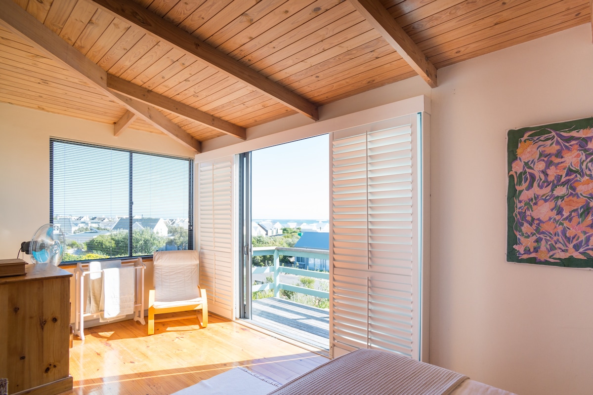 A bright bedroom features wooden beams on the ceiling, with large windows offering a view of the sea. A comfortable chair sits next to the window, and a piece of colorful artwork adorns the wall. Light wood flooring complements the airy atmosphere.