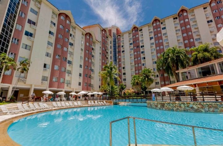 A large swimming pool is surrounded by well-placed lounge chairs under white umbrellas. Palm trees and greenery are visible in the background, while an expansive building with pink and beige features complements the inviting atmosphere of the area.
