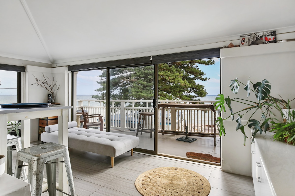 Bright living space features large glass doors leading to a private balcony, offering ocean views. A cozy sofa is positioned nearby, complemented by natural light from the windows. Greenery from potted plants adds a touch of nature, enhancing the airy atmosphere.