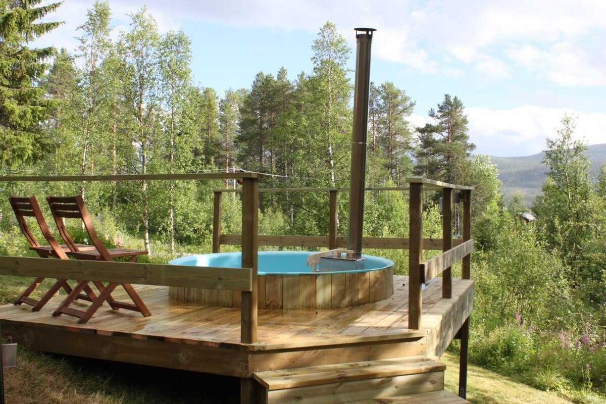 A wooden deck features a circular, heated outdoor bath surrounded by lush greenery. Two wooden lounge chairs are positioned nearby, offering a serene view of the surrounding trees and distant hills.