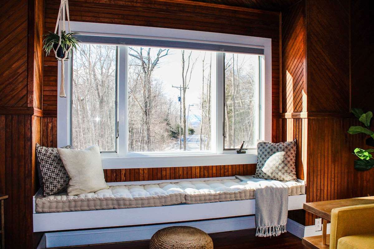 A cozy window nook features a cushioned seat adorned with pillows, offering a serene view of the surrounding trees. Natural light fills the space, highlighting the warm wooden accents and inviting atmosphere. A soft throw blanket is draped over the side.