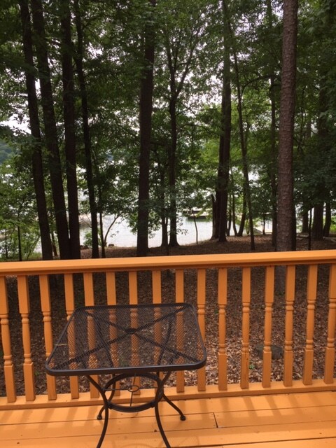 A wooden deck area is visible, featuring a round metal table set among trees. The surrounding foliage provides a natural backdrop, with glimpses of Lake Hartwell visible through the trees in the distance.