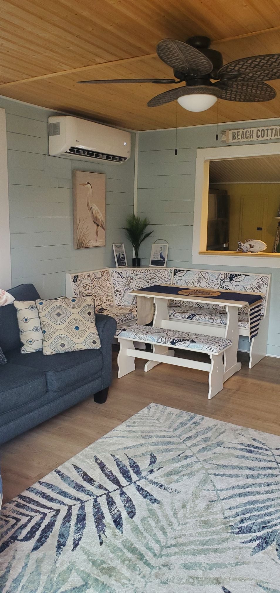 A cozy living area is furnished with a dark sofa and a patterned seating bench. Decorative pillows enhance the seating. A ceiling fan hangs above, and light wood paneling complements the soft-colored walls. A tropical-themed area rug adds warmth and texture to the space.