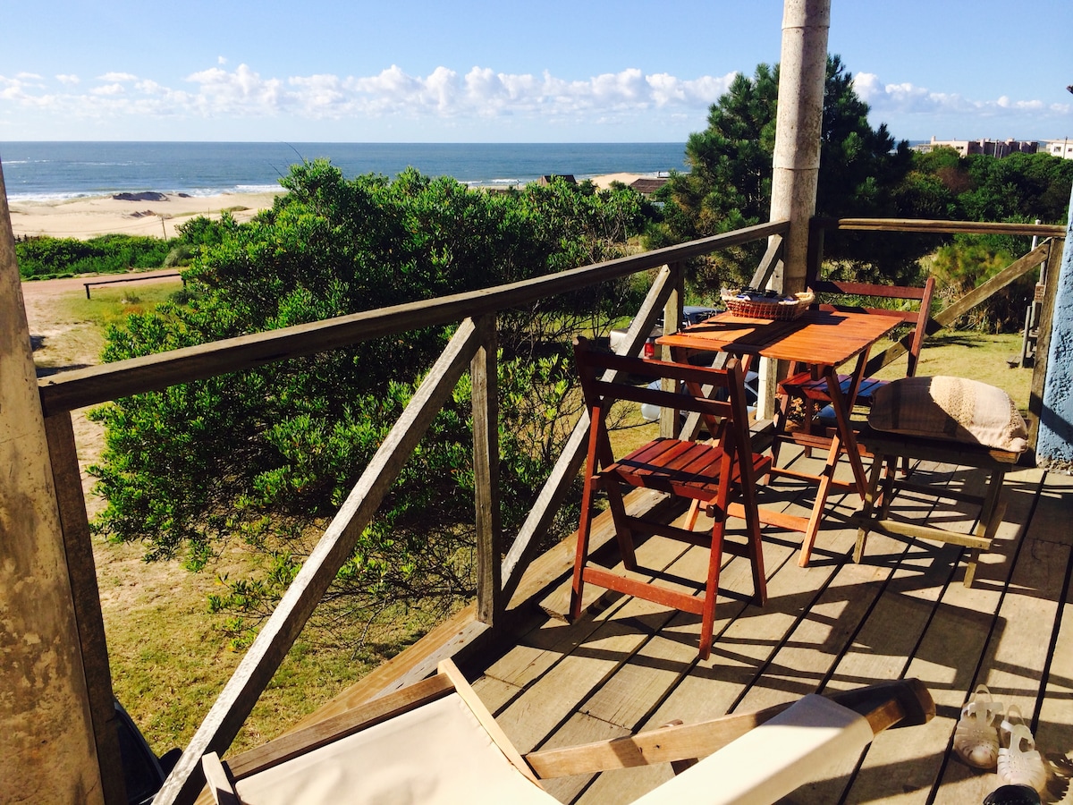 A wooden deck features a view of the ocean, bordered by greenery. Two folding chairs and a small table occupy the space, inviting relaxation. The sky is clear with scattered clouds, and the beach is visible in the distance.