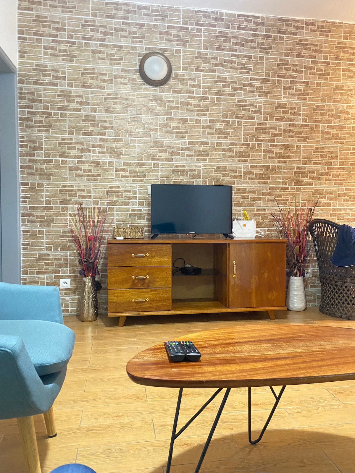 A living room space is shown, featuring a wooden coffee table and a television positioned on a mid-century style console. Textured wallpaper in warm tones provides a backdrop, complemented by decorative plants in stylish vases and a comfortable blue chair.