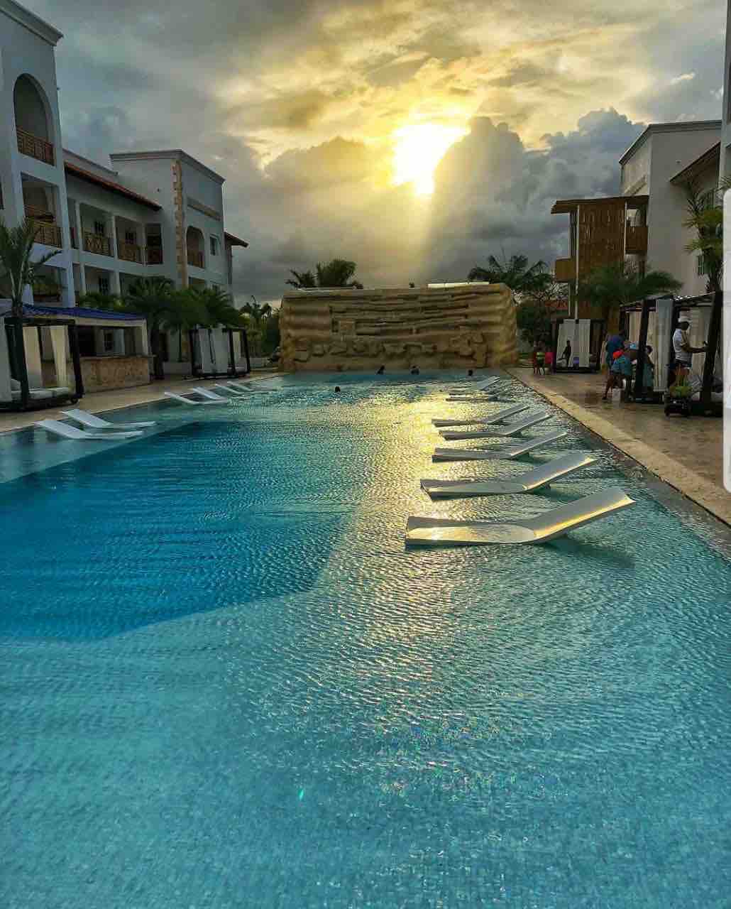 A tranquil swimming pool reflects the soft glow of the sun setting behind a clouded sky. Lounge chairs are arranged along the water's edge, inviting relaxation. Lush palm trees frame the pool area, enhancing the serene environment.