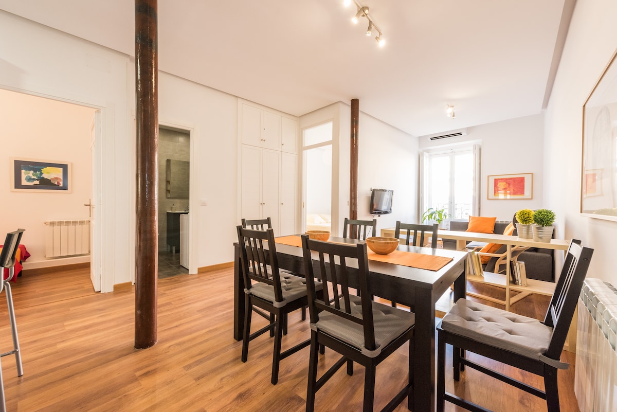 A spacious dining area is featured with a long wooden table surrounded by eight black chairs. Natural light fills the space through large windows, enhancing the warm wood flooring. A small couch and decorative plants create a welcoming atmosphere in the corner of the room.