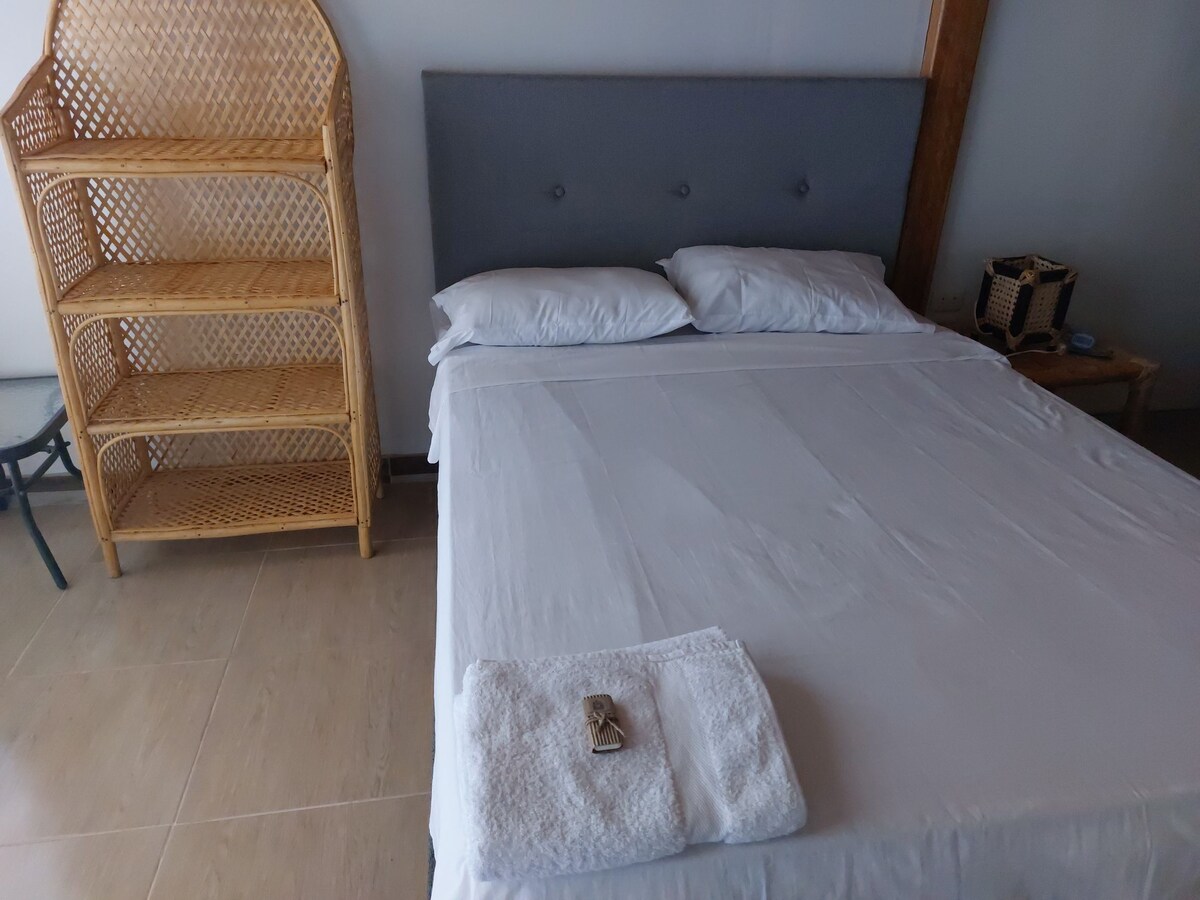 A cozy sleeping area features a spacious bed with crisp white linens. A neatly folded towel is placed at the foot of the bed. Woven storage shelves and a small side table are visible, providing functional decor in the calming room.