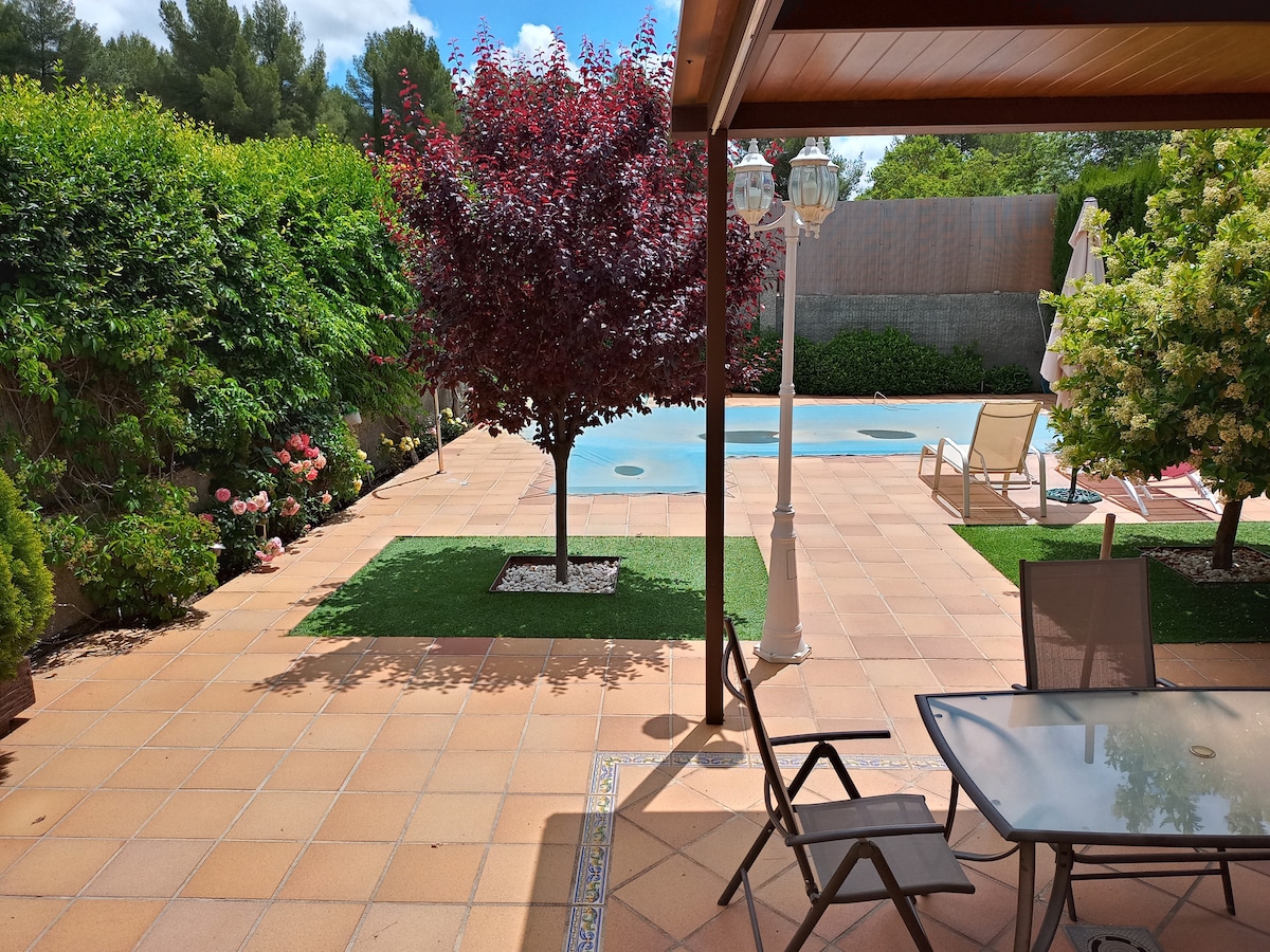 A spacious patio is seen, featuring a dining table with chairs and a pool in the background. Lush greenery and a decorative tree enhance the setting. The area provides a peaceful environment with well-maintained landscaping and clear skies above.