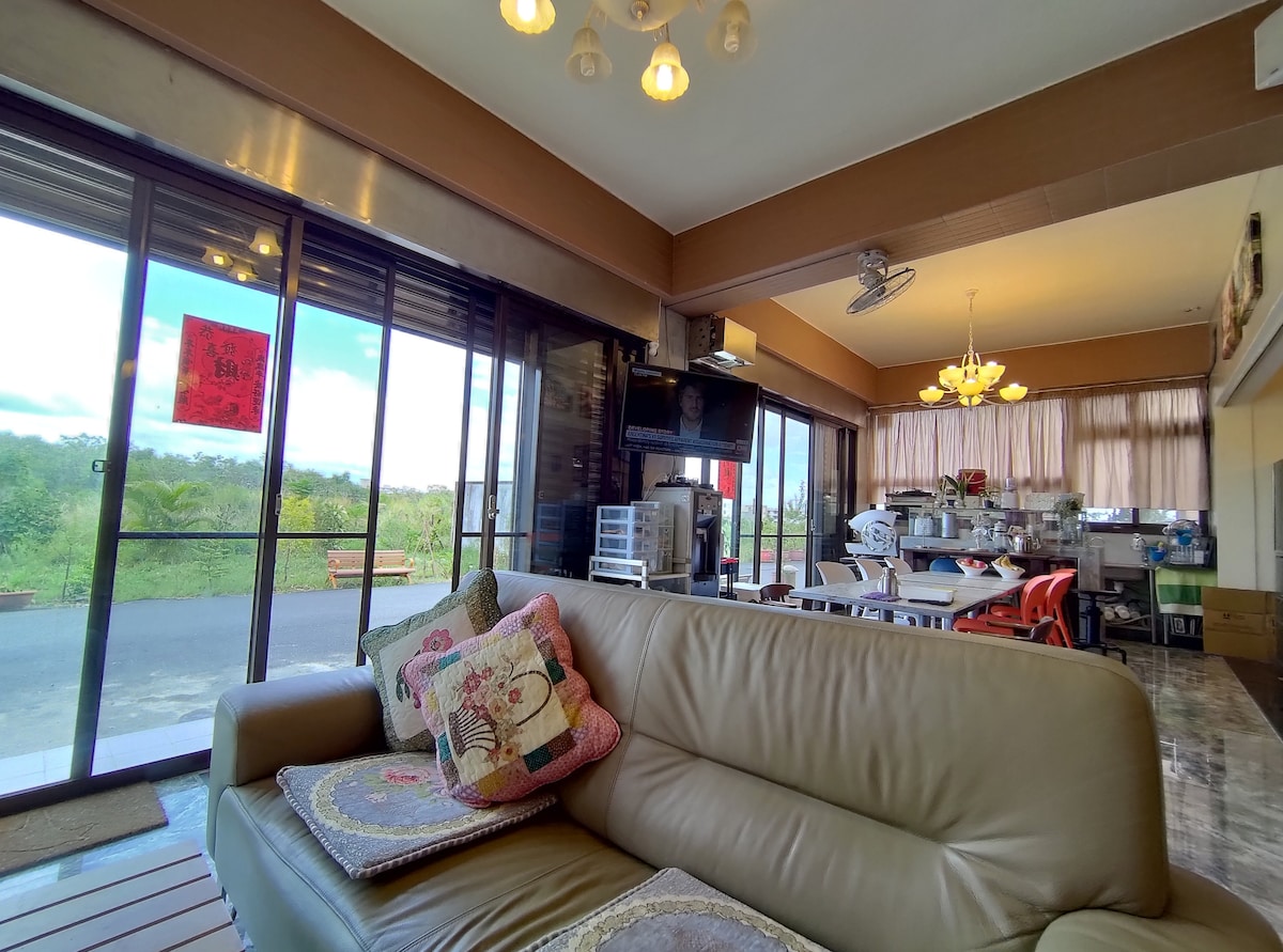 A spacious living area features a light-colored sofa adorned with decorative cushions. Large glass doors provide views of the surrounding greenery, while a dining space with colorful chairs is visible in the background. Soft lighting enhances the relaxed ambiance.