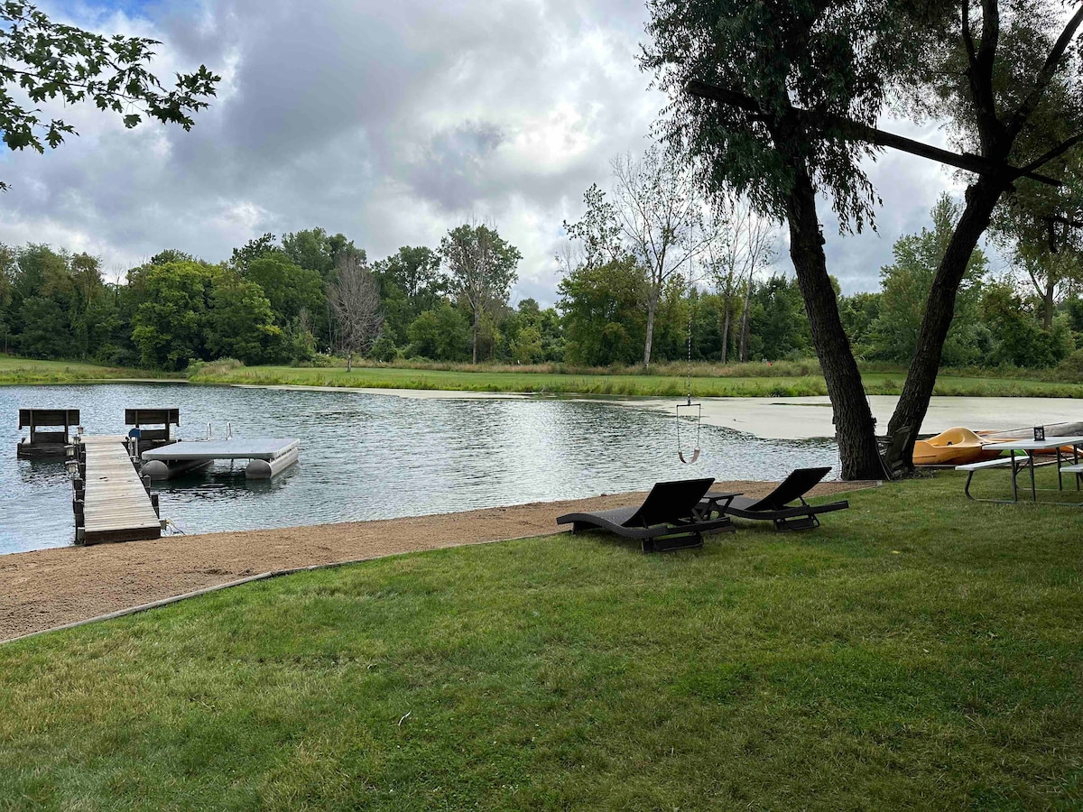 A serene view of a pond is featured, with two lounge chairs positioned on the grassy shore. A wooden dock extends into the water, while a kayak rests on the bank nearby. Lush greenery frames the scene, suggesting a peaceful natural environment.