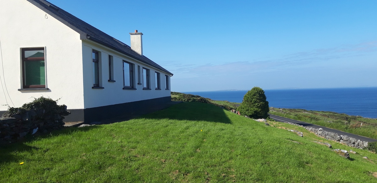 A detached house is positioned near the edge of a gently sloping green lawn. The exterior features white walls and dark window frames. The expansive view of the sea and distant coastline stretches into the horizon under a clear blue sky.