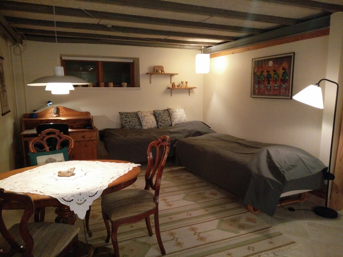 A cozy room features two spacious beds covered with grey bedspreads, positioned alongside a small writing desk and a wooden chair. A dining table is set with light-colored linens. Decorative shelves hold various items above, while gentle lighting creates a welcoming space.
