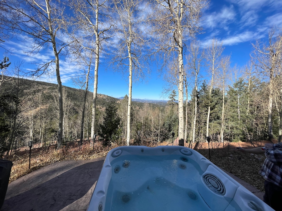 A spacious hot tub is positioned on a paved area, with clear views of the surrounding forest and mountains. Tall aspen trees rise nearby, framing the landscape under a bright blue sky. The serene outdoor setting invites relaxation and connection with nature.