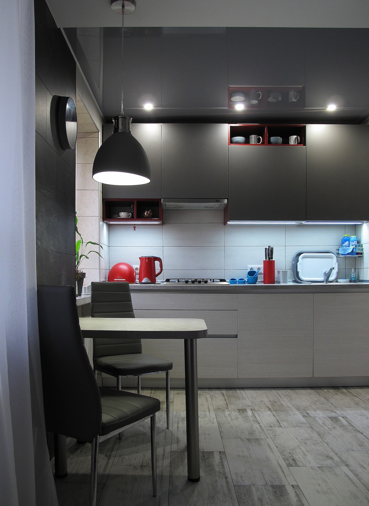 The kitchen area displays modern cabinetry with integrated lighting, featuring a compact table and two chairs. A kettle and various utensils are visible on the countertop, and open shelving showcases a selection of dishware. Stylish pendant lighting hangs above the dining space.