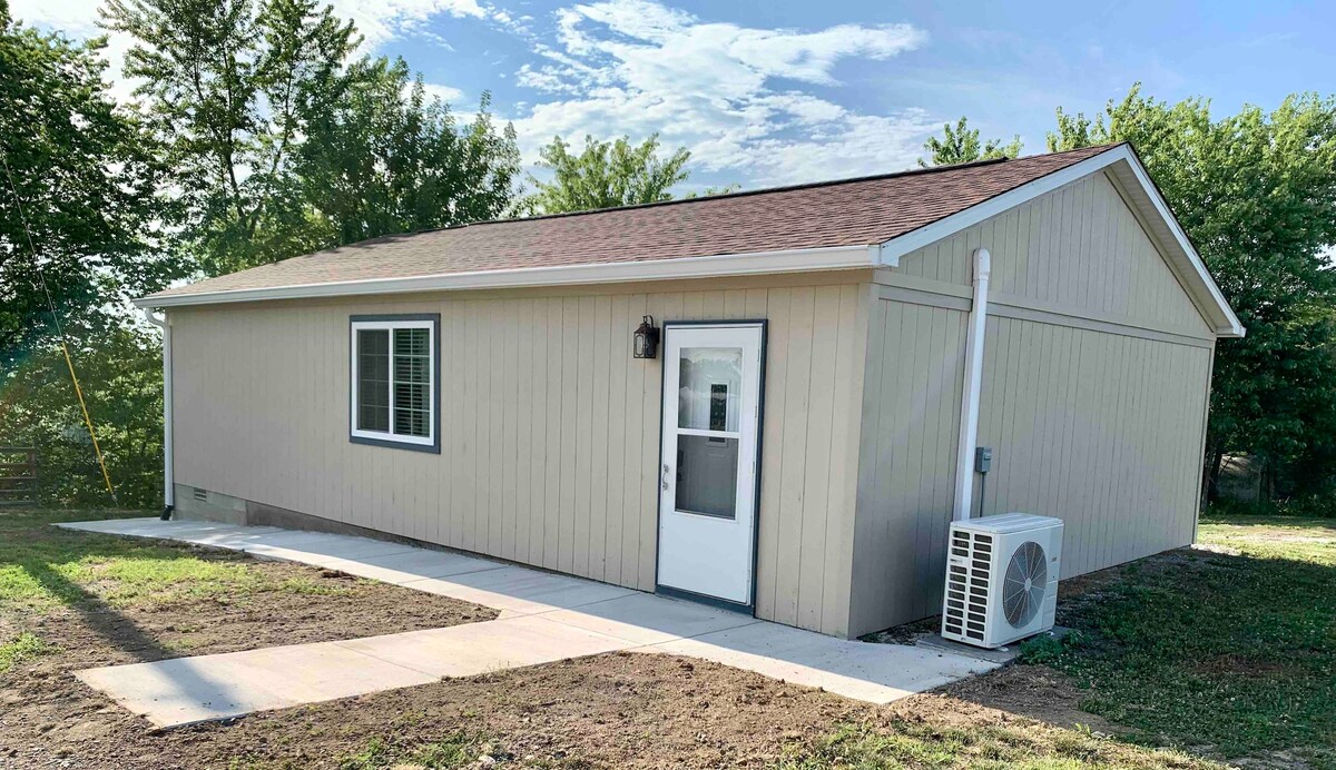 A single-story cabin is shown surrounded by greenery, featuring beige siding and a sloped roof. A large window provides natural light to the front room, and a white door is positioned centrally. An air conditioning unit is attached to the exterior wall.