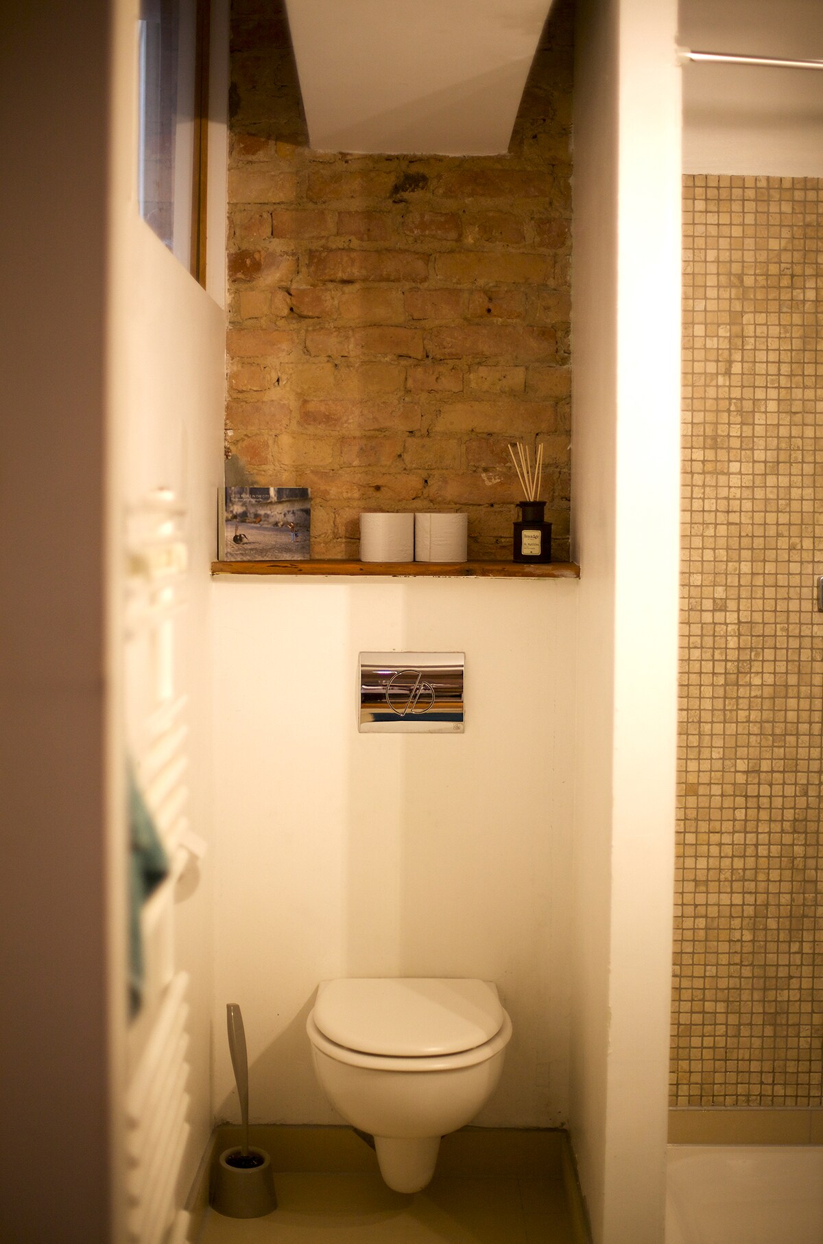 The bathroom features a compact design with exposed brick walls. A modern toilet is positioned beside a glass shower, while a shelf displays decorative elements and toiletries. Soft lighting enhances the ambiance, creating a functional yet inviting space.