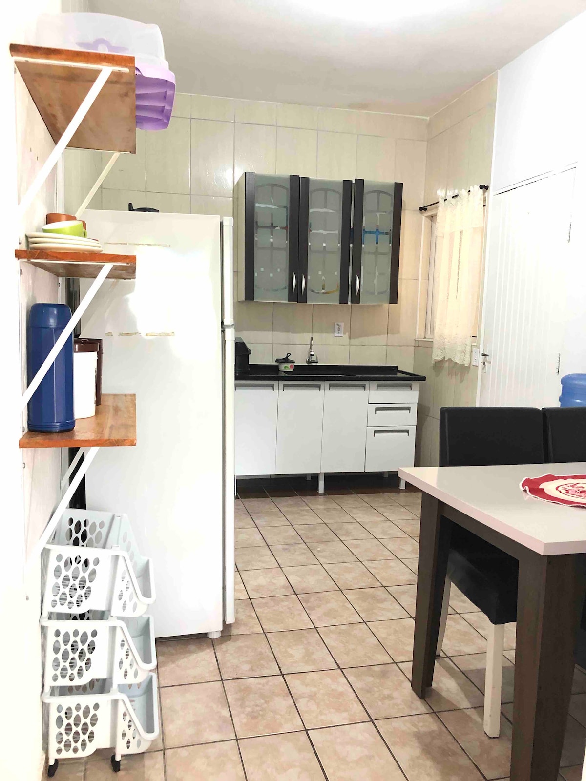 The kitchen is equipped with modern cabinetry, a refrigerator, and a countertop area. A dining table with four chairs is present, along with open shelving displaying various kitchen items. Soft natural light filters through a nearby window, illuminating the tiled floor.