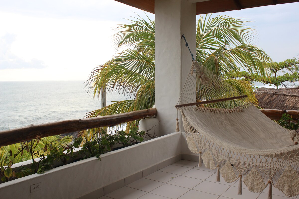 A macrame hammock is suspended from wooden beams on a spacious balcony, overlooking the tranquil sea. Lush palm trees frame the scene, with soft ocean waves visible in the distance under an expansive sky.