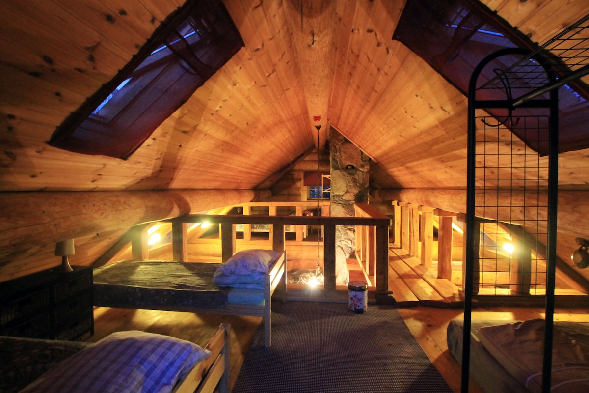 The sleeping area features a wooden ceiling with exposed beams, softly lit by warm lights. Two smaller beds are positioned against the walls, while a cozy nook with a view from the skylights enhances the inviting atmosphere. A peaceful space for restful nights.