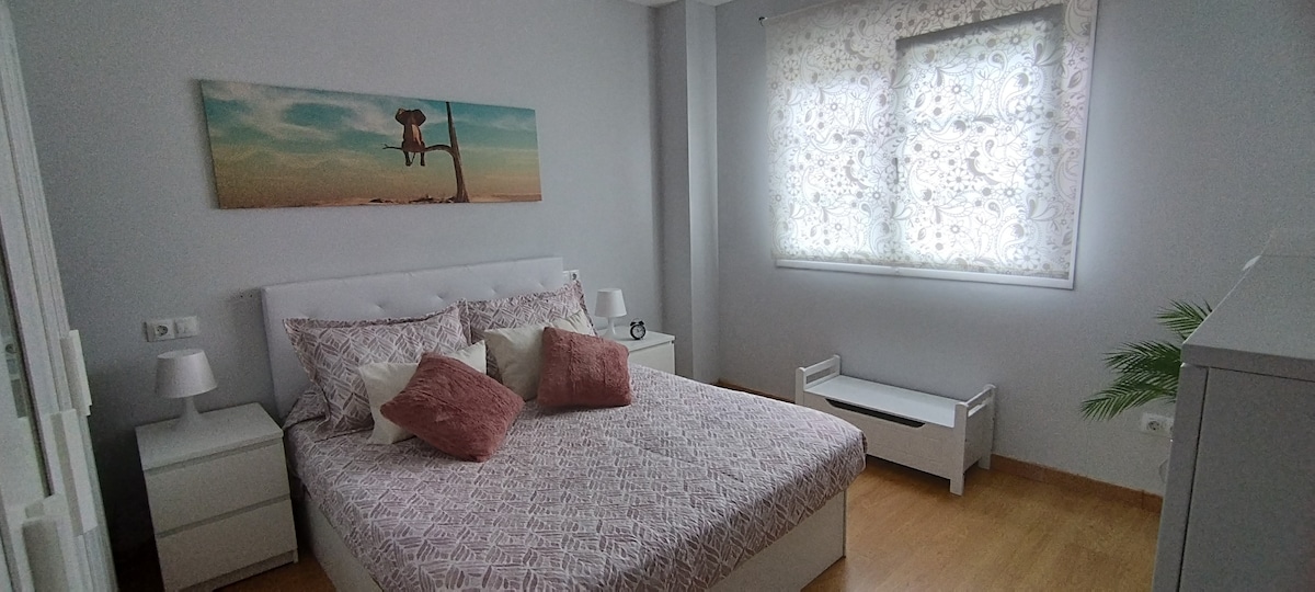 A bright bedroom features a spacious bed adorned with patterned linens and decorative cushions. Two nightstands with lamps flank the bed, while a window with sheer curtains allows natural light to fill the room. A small bench and a plant add a touch of greenery.