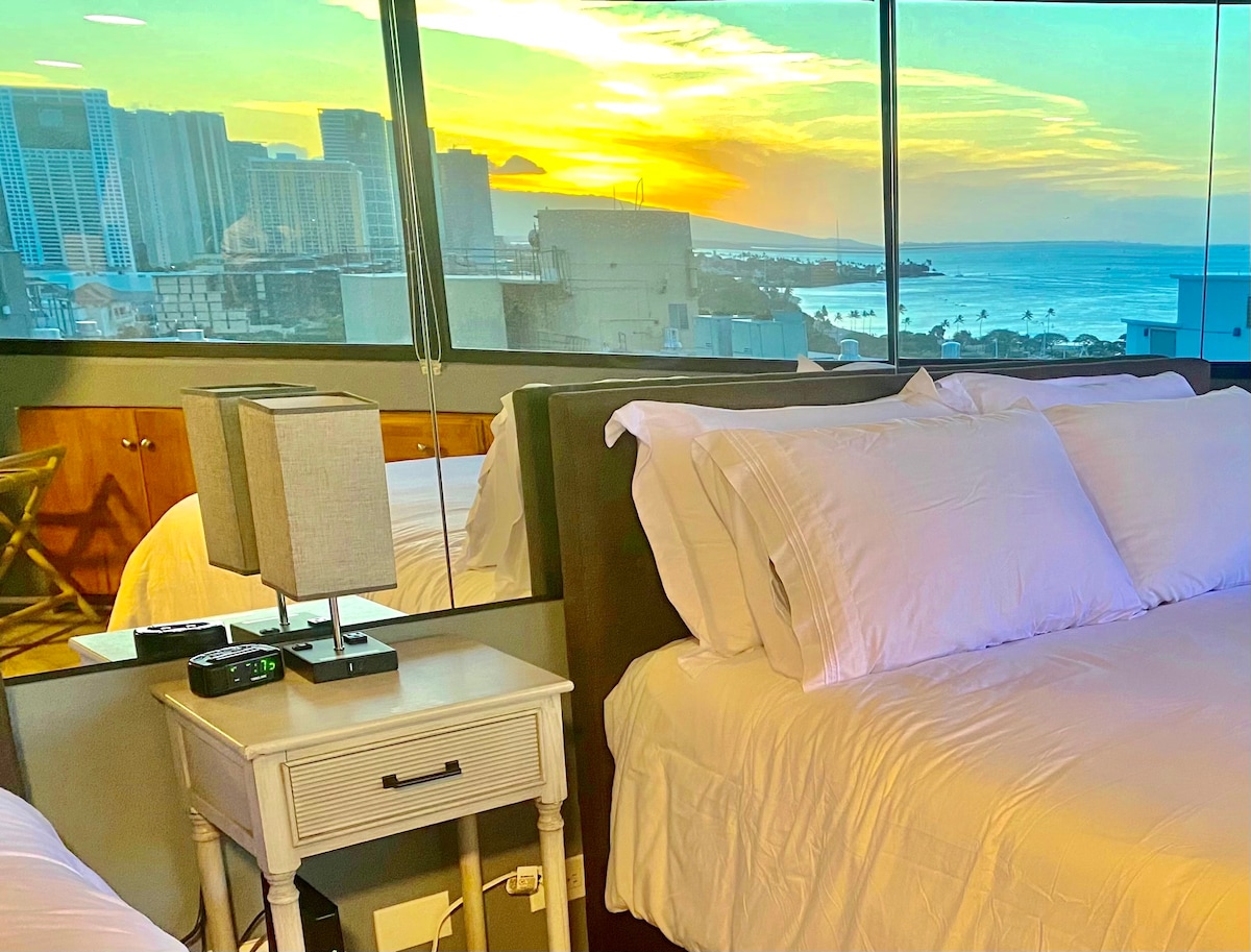 A spacious sleeping area features two inviting beds with clean white linens. A bedside table holds a lamp and a clock. In the background, large windows showcase a vibrant sunset over the ocean and city skyline, creating a serene and calming view.
