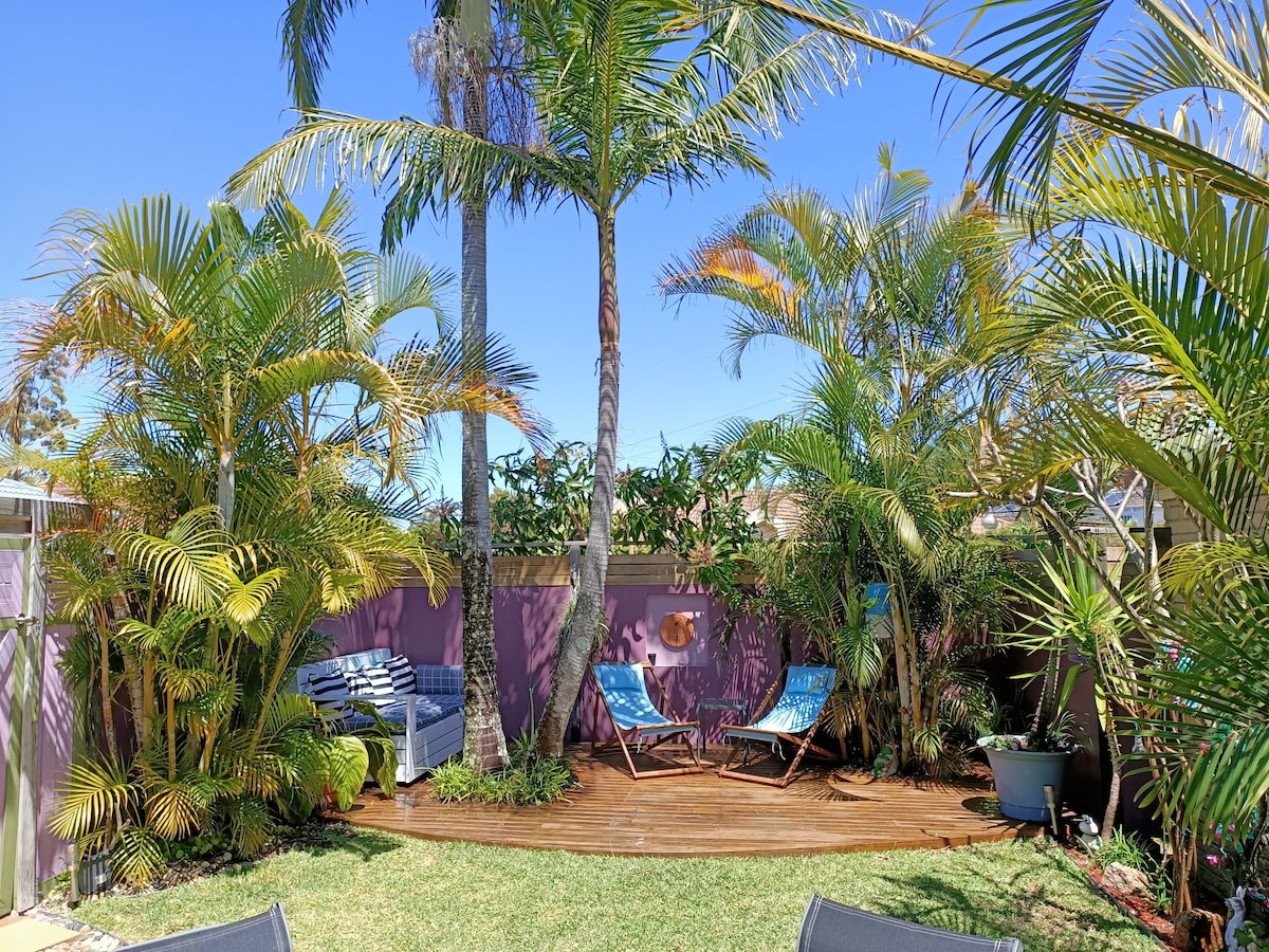 A private courtyard features lush tropical plants, creating a serene atmosphere. Two lounge chairs are positioned on a wooden deck, inviting relaxation. A comfortable seating area is visible, enhancing the outdoor space's welcoming charm, complemented by a vibrant purple wall.