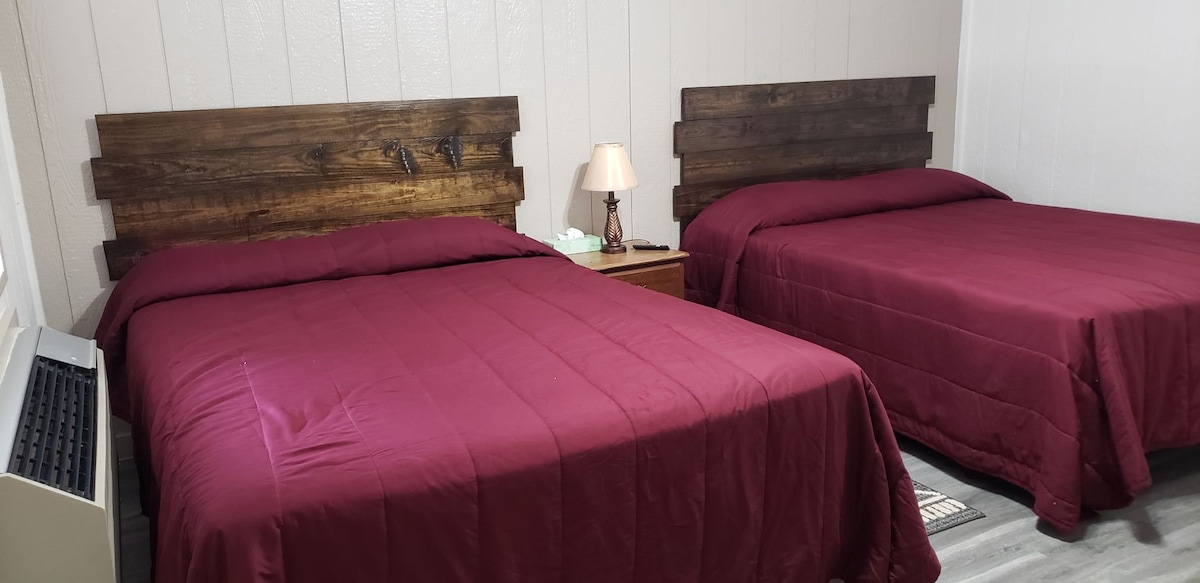 A guest room is presented with two Queen beds, both topped with deep red bedspreads. A small bedside table with a lamp sits between the beds, while a window allows natural light into the space. Air conditioning is available for comfort.
