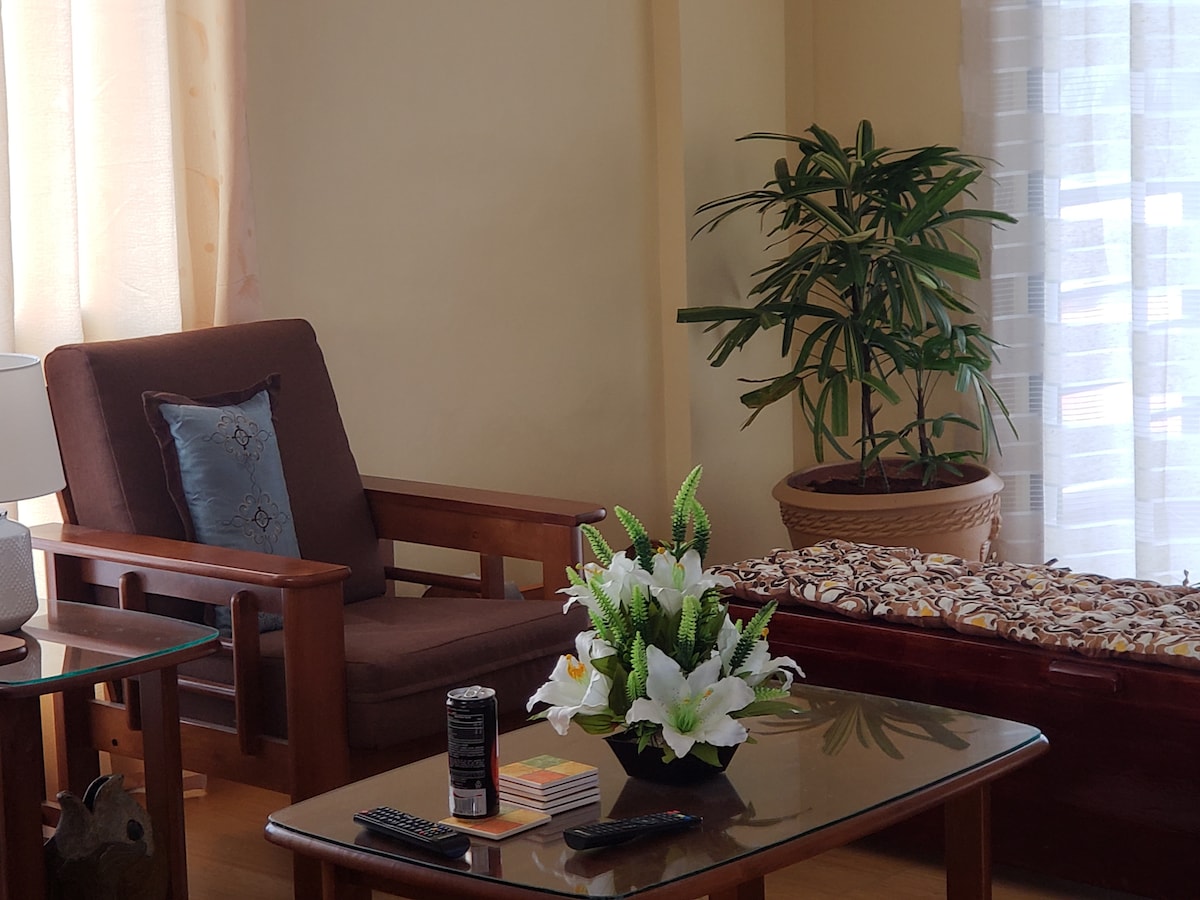 A comfortable seating area is displayed, featuring a wooden armchair and a long bench covered with patterned fabric. A glass coffee table is set with a decorative arrangement of flowers and magazines. Natural light filters through sheer curtains, illuminating the space.