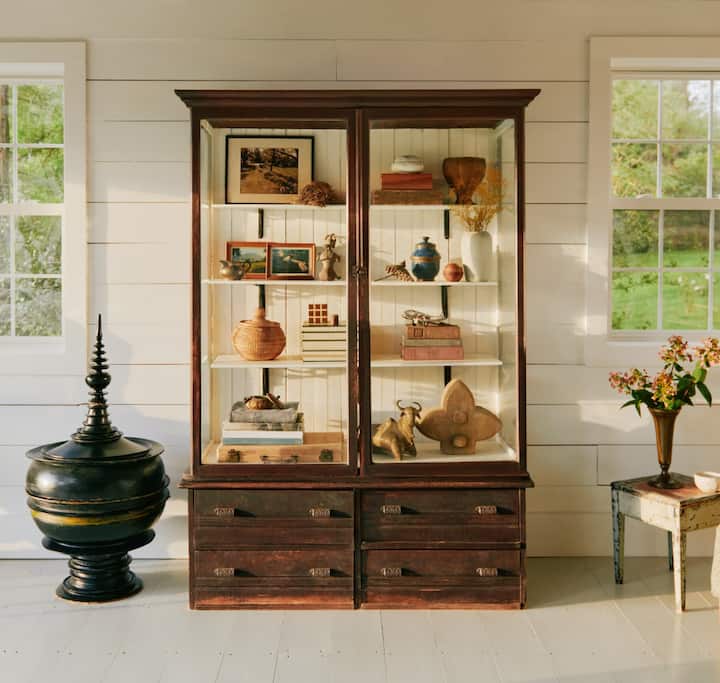 Una foto muestra una sala grande, blanca, con una colección de muebles antiguos que incluye un estante grande de madera y vidrio lleno de una variedad de objetos vintage de valor.
