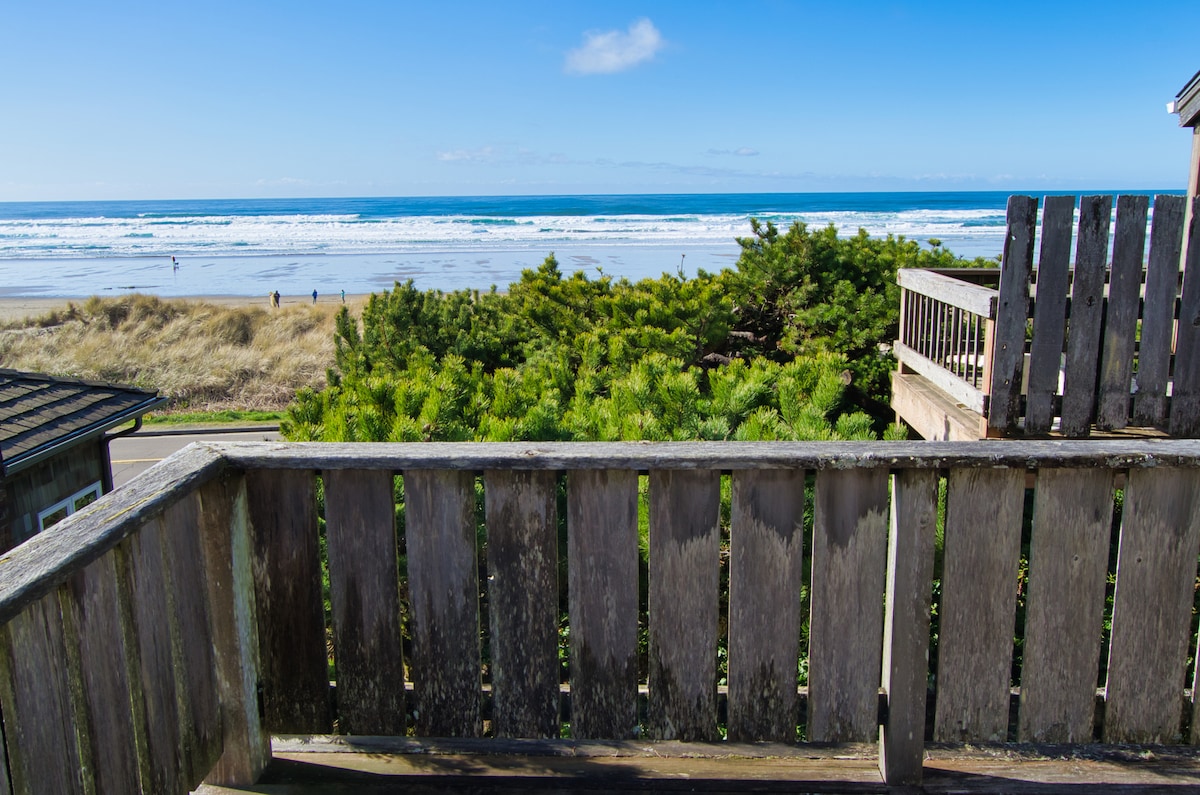 A spacious deck is visible, showcasing a wooden railing and a panoramic view of the Pacific Ocean and sandy beach. The gentle waves can be seen lapping at the shore, with people walking along the beach in the distance. Lush coastal vegetation frames the scene.
